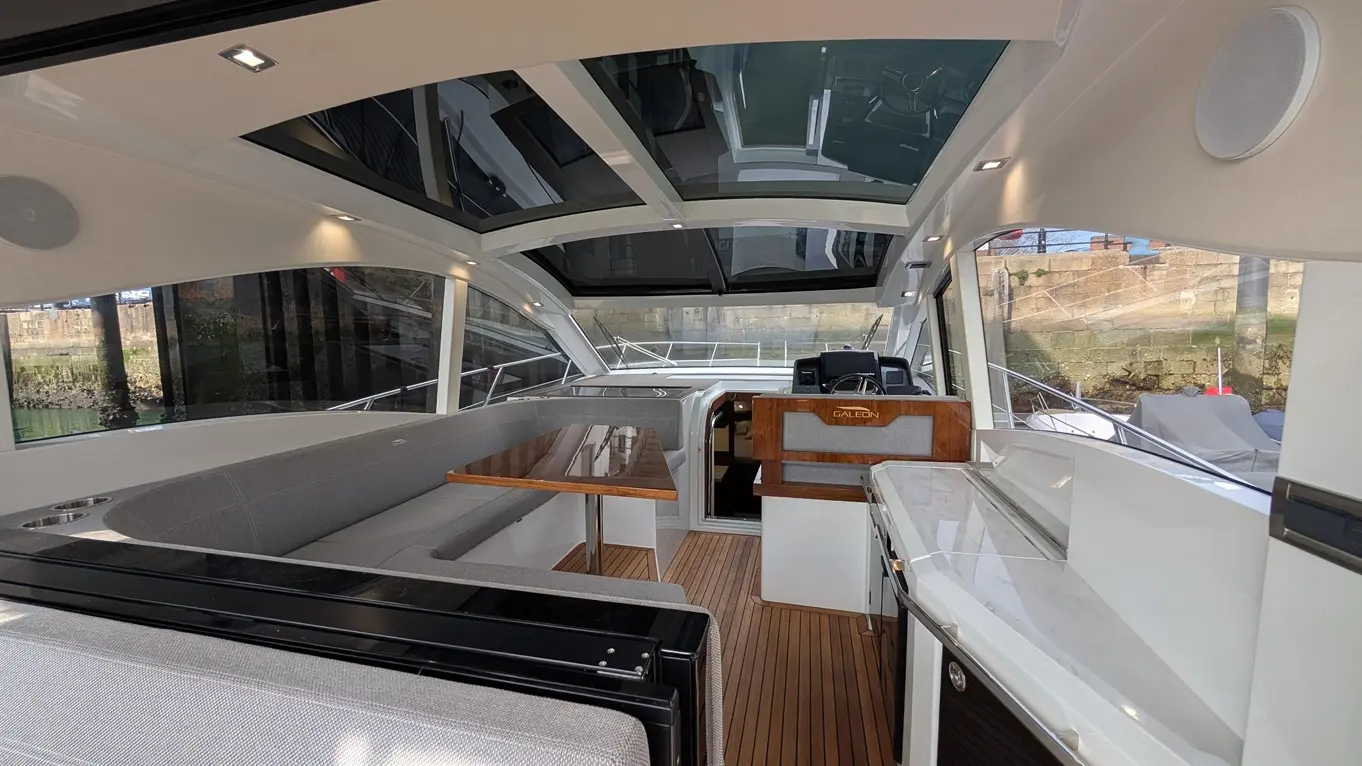 Modern boat interior with a dining area, helm station, and large windows, showcasing wooden and grey furnishings.